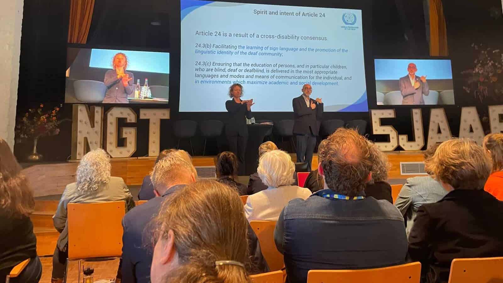A conference audience listens to presenters on stage with two sign language interpreters; a slide about Article 24 and language rights is displayed on the screen.