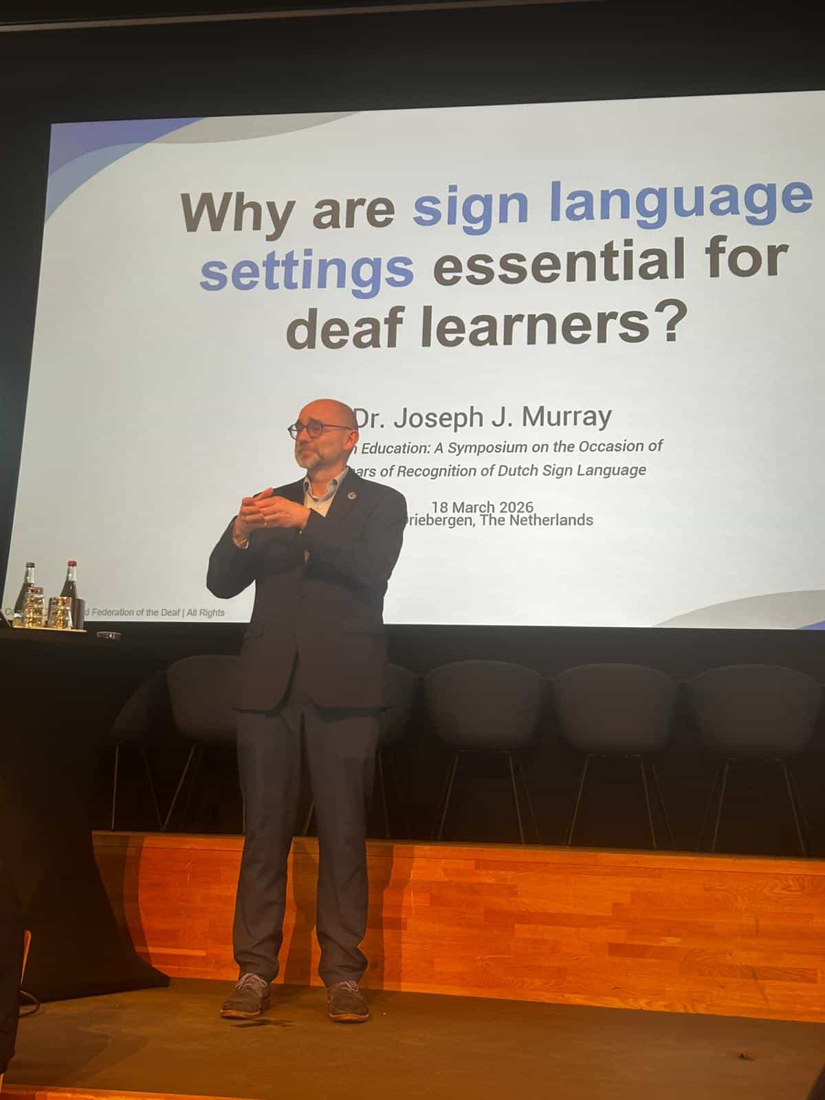 A man stands on stage signing in front of a presentation slide titled "Why are sign language settings essential for deaf learners?" at a symposium on Dutch Sign Language.
