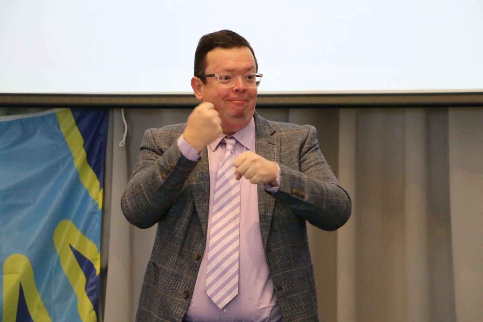 A man in a suit and striped tie is using sign language in front of a blue and yellow banner and a projector screen.