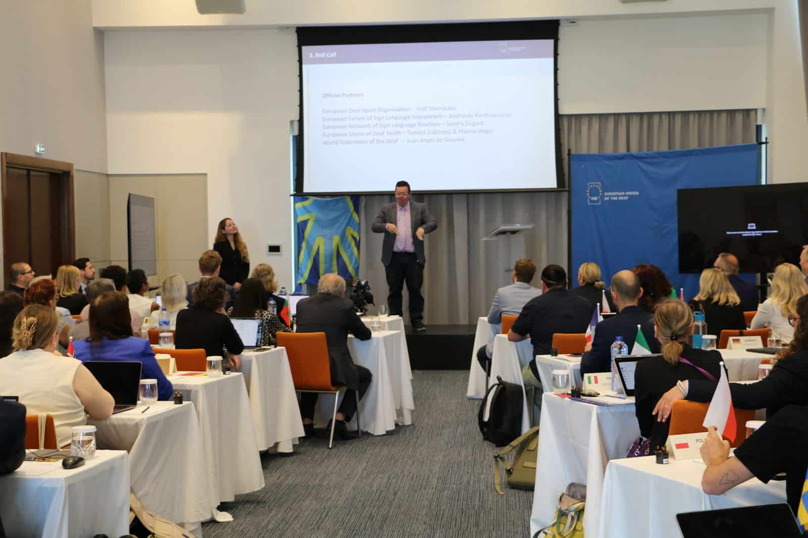 A man in a suit speaks at a conference in a room with seated attendees, presentation slides, and flags visible on tables.