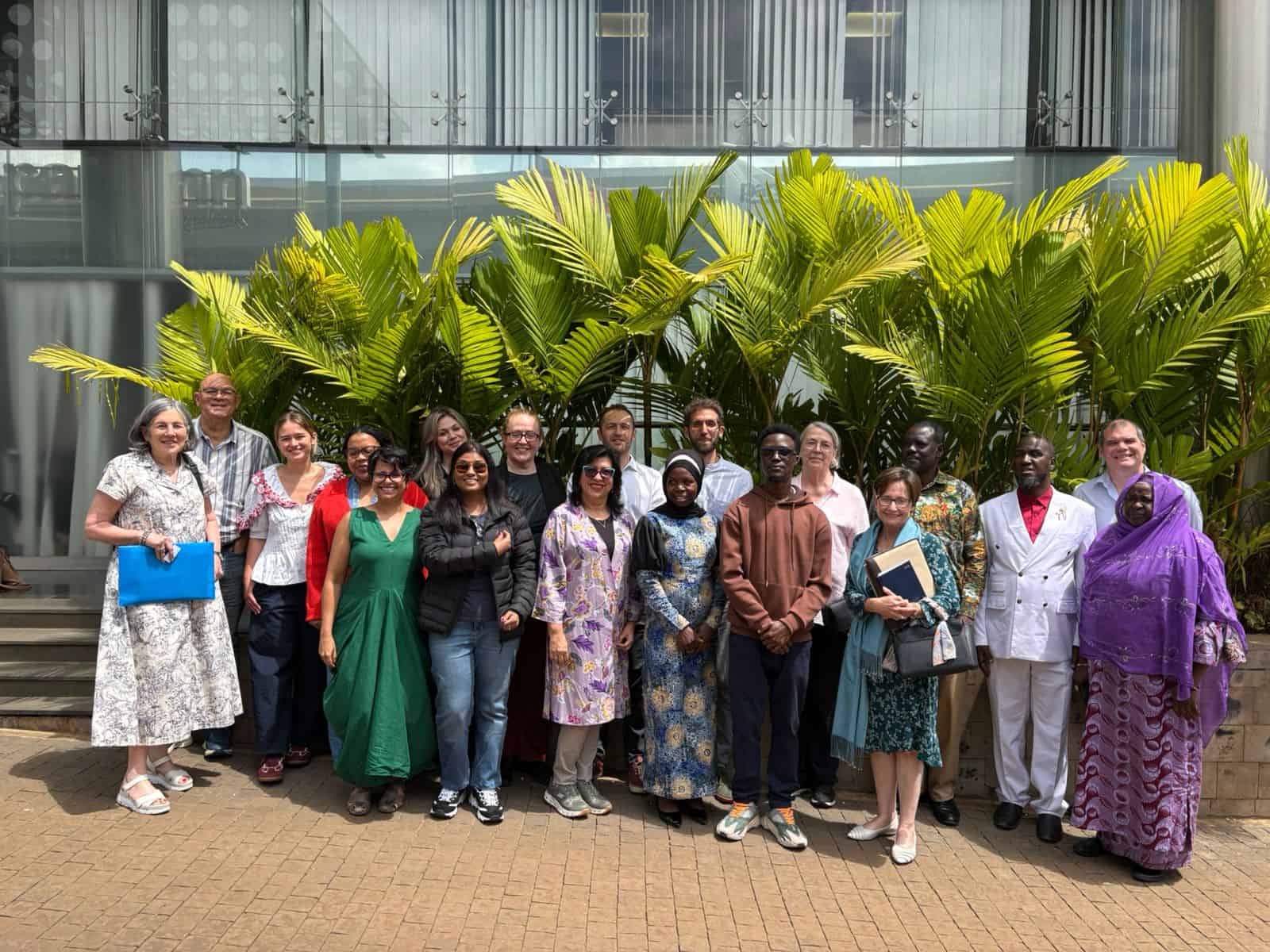 A group of people pose for a photo in front of green plants and a modern building, standing on a paved area in daylight.