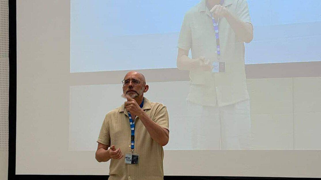 A man stands on a tiled floor speaking into a microphone in front of a large screen displaying his live image.