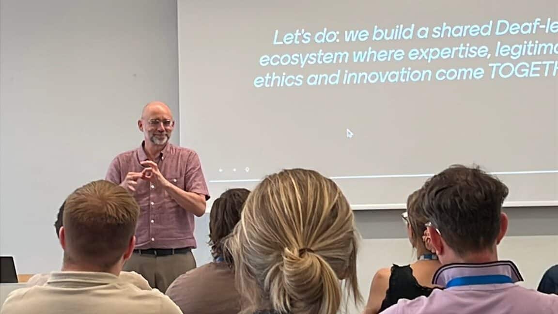 A speaker stands at the front of a classroom addressing an audience, with a presentation slide behind him that discusses building a shared Deaf-led ecosystem.