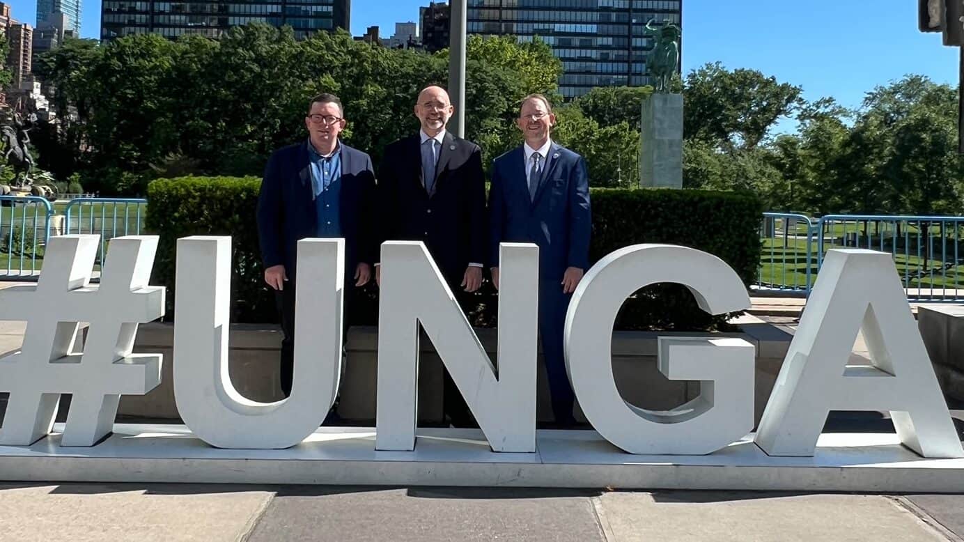 Three men in suits stand behind a large "#UNGA" sign outdoors, with tall modern buildings and a clear blue sky in the background.