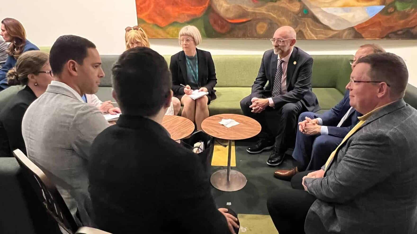 A group of people in business attire sit in a circle having a discussion in a room with green sofas and abstract art on the wall.