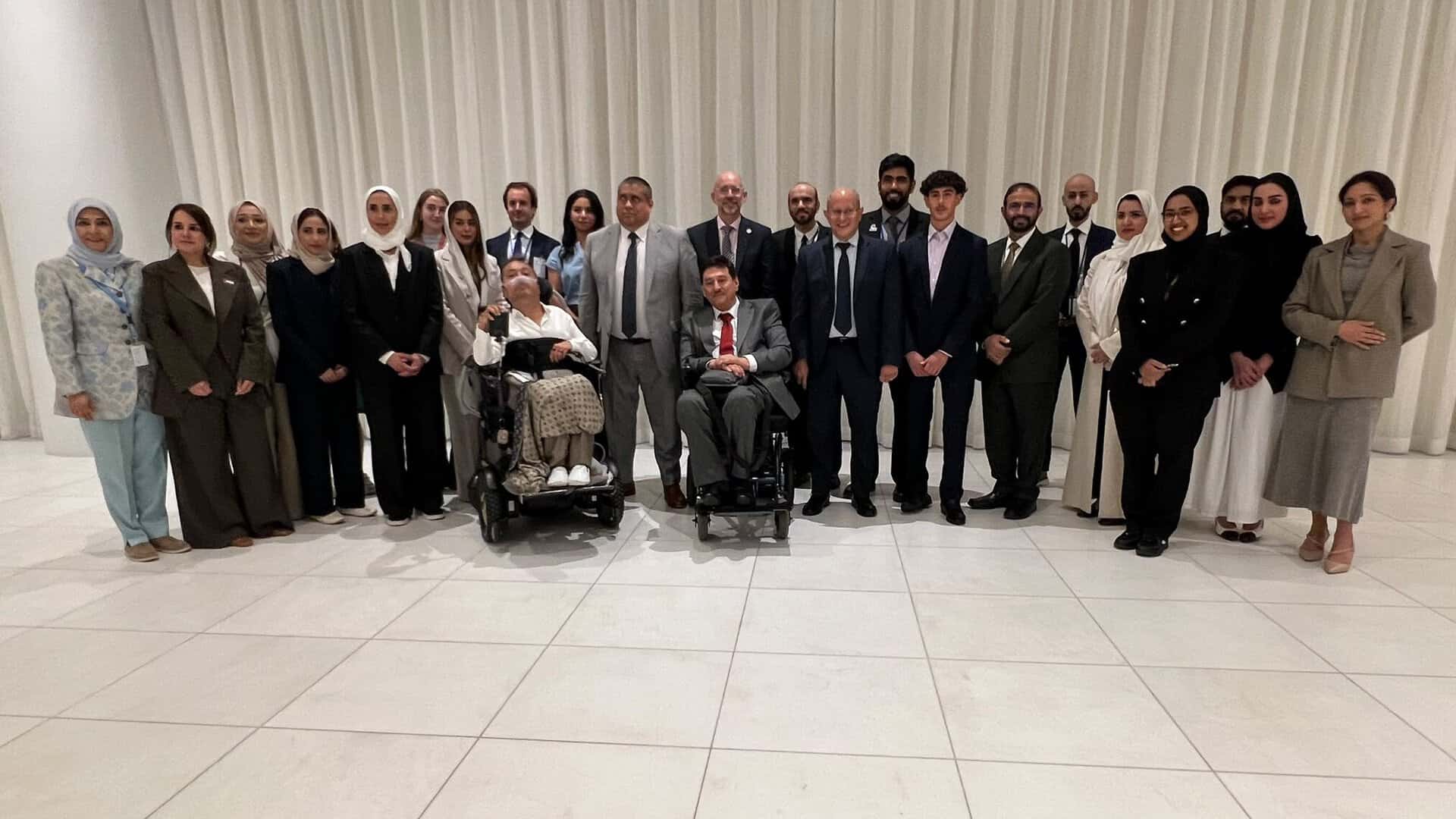 A group of 21 people, including two in wheelchairs, pose together indoors against a white curtain backdrop.