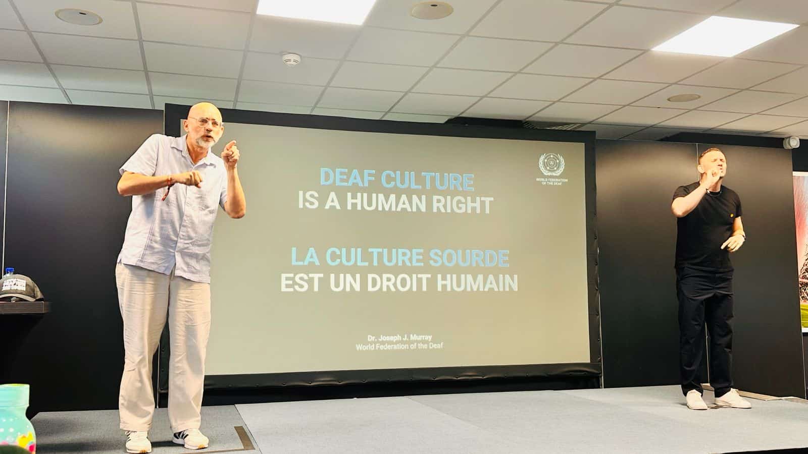 Two presenters stand on a stage; behind them, a screen displays the text "Deaf culture is a human right" in English and French at an event.
