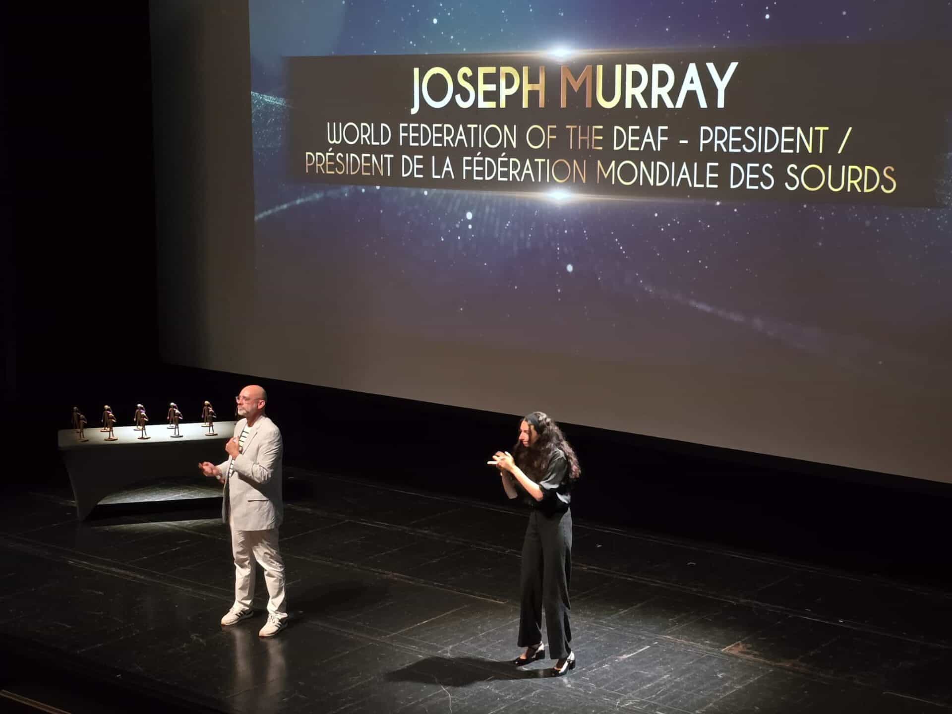 Two people stand on a stage, one signing and one speaking, in front of a screen showing "Joseph Murray, World Federation of the Deaf – President." A table with awards is visible.