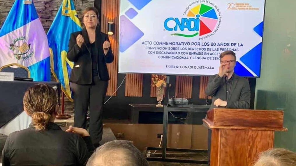 A woman signs in front of an audience next to a man seated at a desk, with Guatemalan flags and a presentation screen displaying the CNADI logo and text in Spanish.