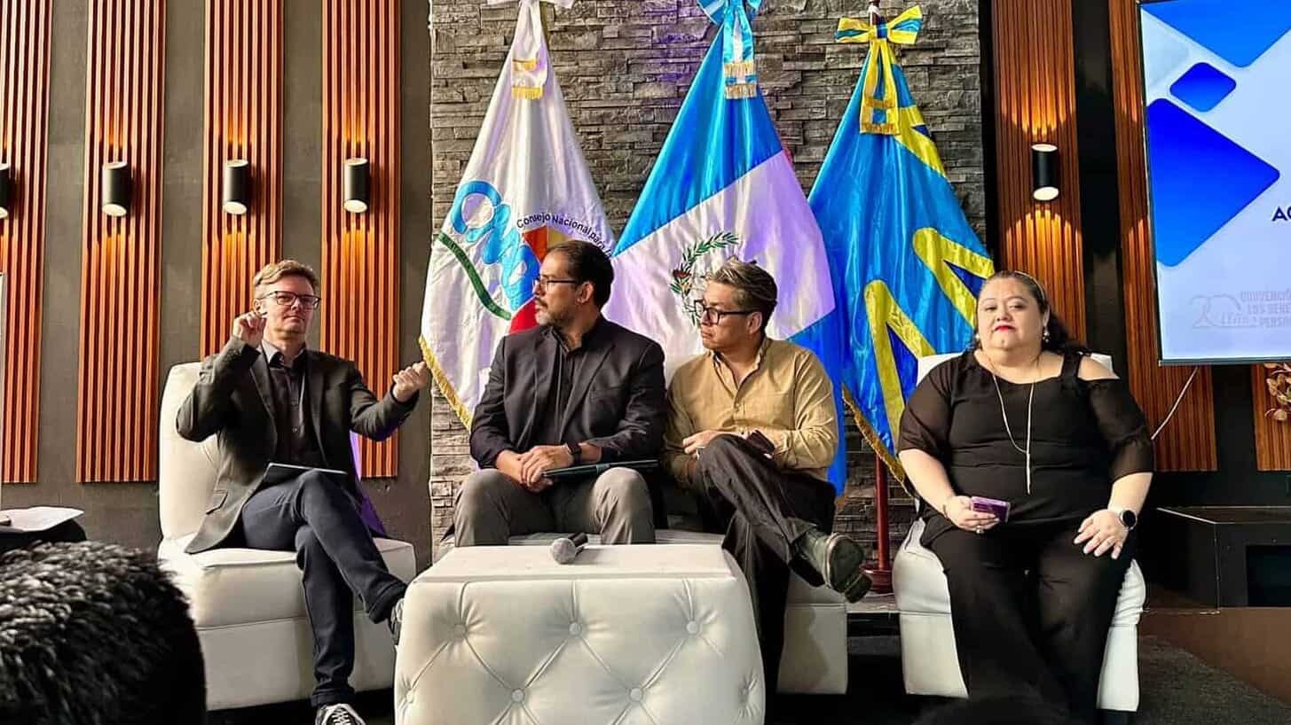 Five people sit on a stage in front of flags and a large screen during a formal event; one person is using sign language.