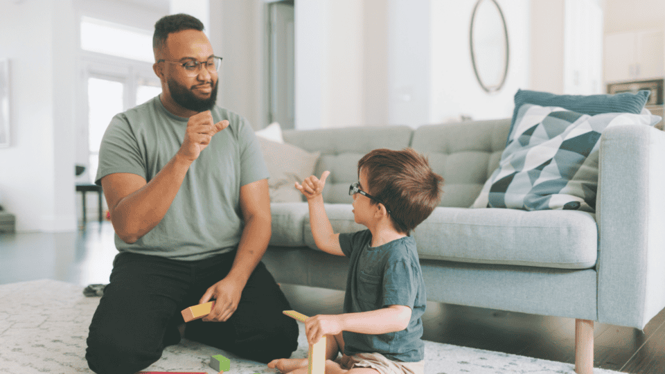 An adult and a child sit on the floor in a living room, wearing glasses and gesturing to each other whilst playing with colourful wooden blocks.