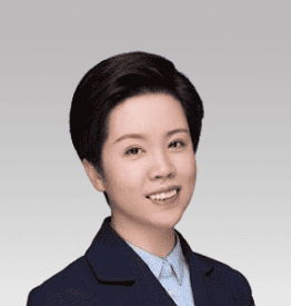 A person with short dark hair smiles, wearing a dark blazer and light blue shirt, set against a light gray background.