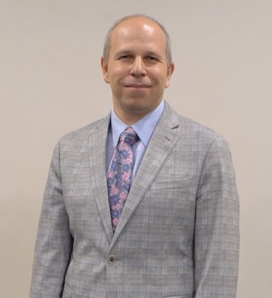 A man in a gray suit with a floral tie standing against a plain background.