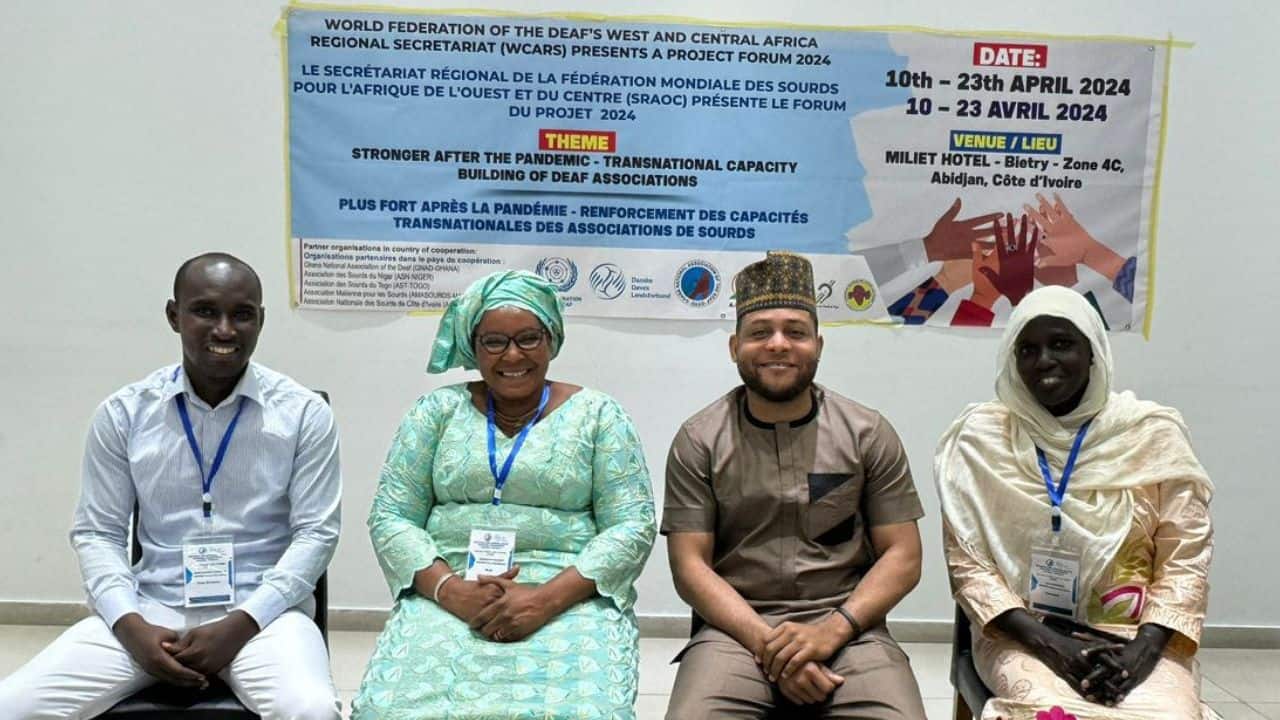 Four people seated in a row, smiling, in front of a bilingual conference poster about the World Federation of the Deaf's Projets Forum 2024 in Abidjan, Côte d'Ivoire.