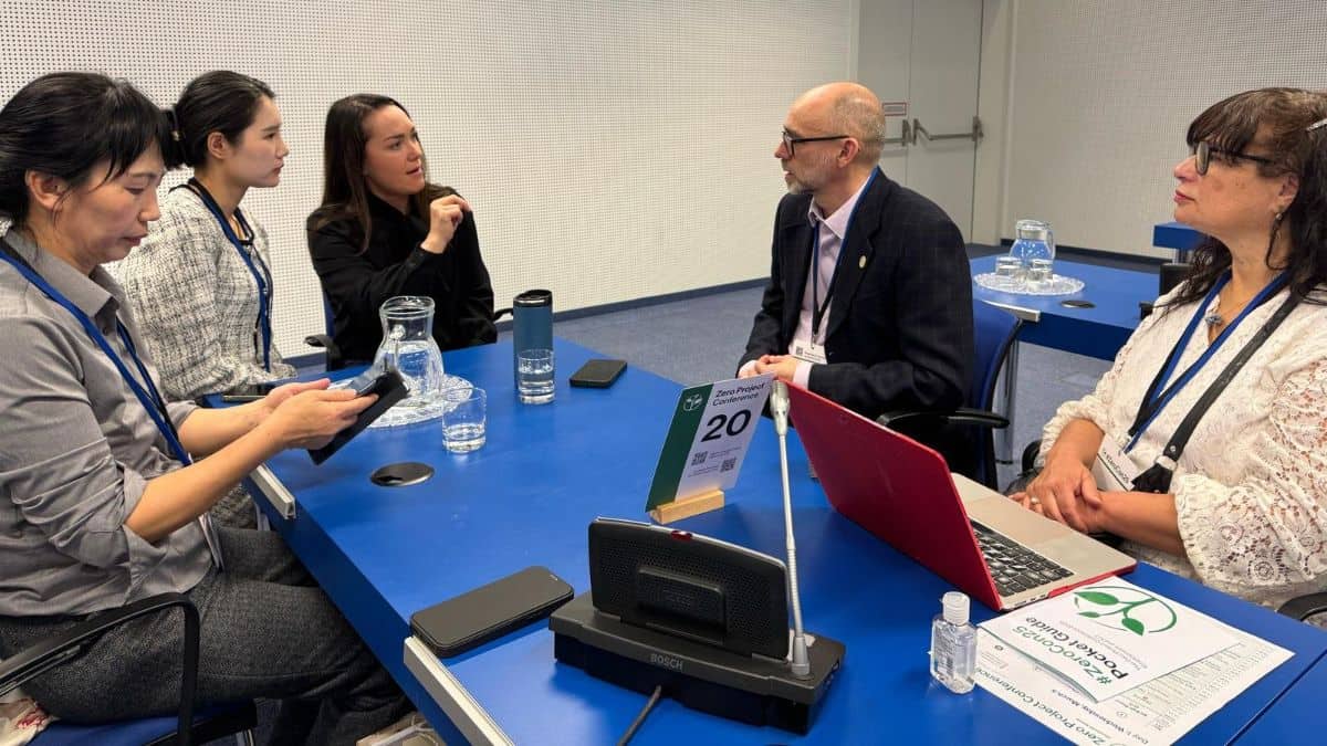 Five people are engaged in discussion around a blue conference table. Papers, a laptop, and a nameplate displaying the number 20 are on the table.