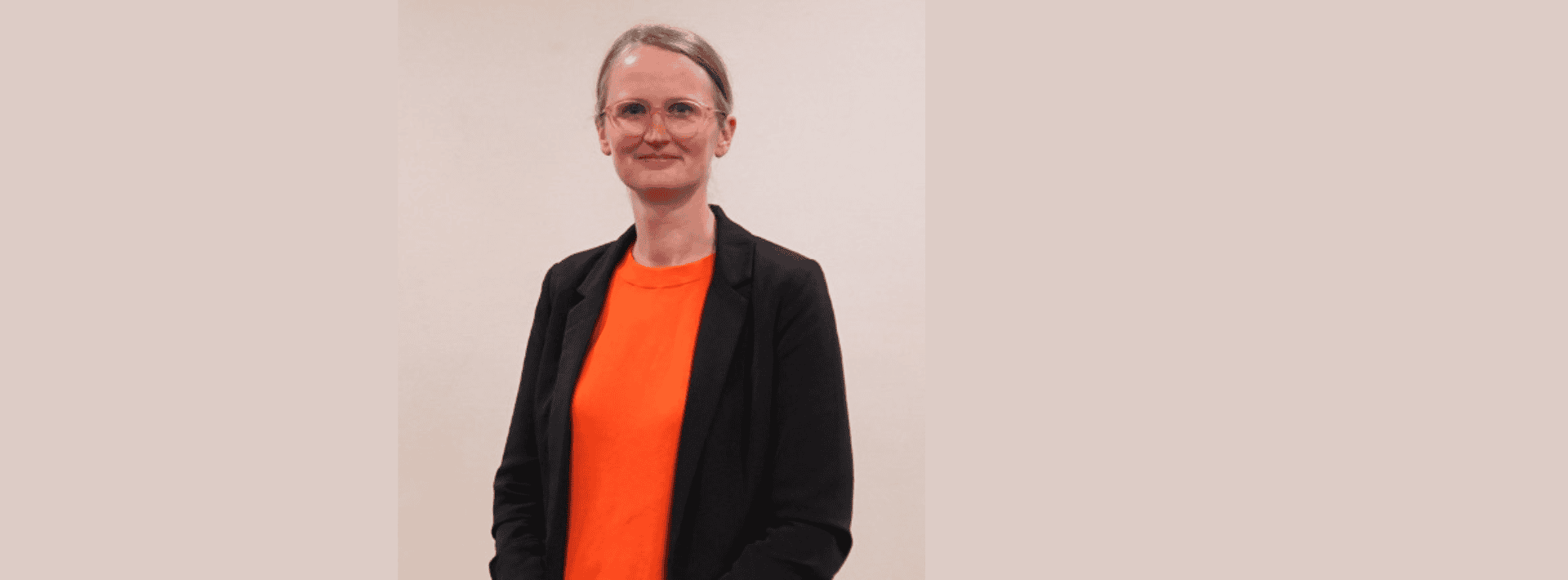 A person with glasses and tied-back hair wears an orange shirt and a black blazer, standing against a plain beige background.