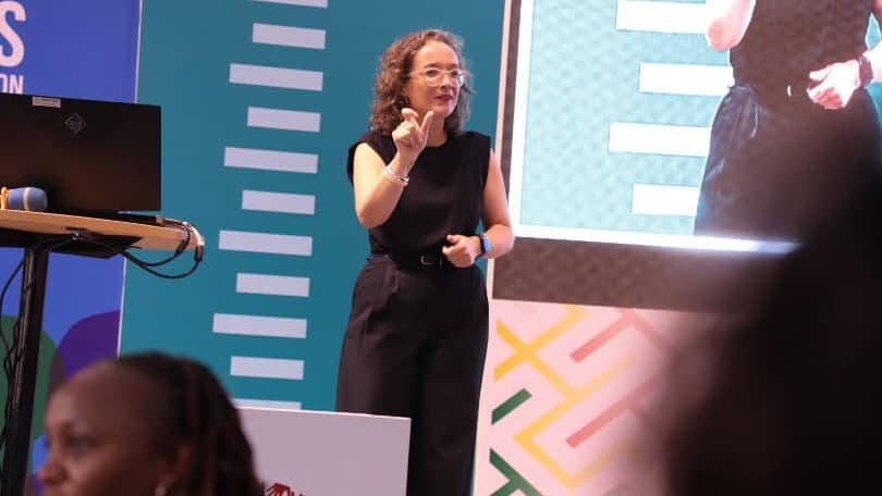 A woman in black speaks and gestures on stage during a presentation, with a large screen and colorful patterned backdrop behind her. Audience members are visible in the foreground.