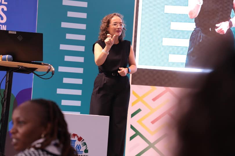 A woman in black speaks and gestures on stage during a presentation, with a large screen and colorful patterned backdrop behind her. Audience members are visible in the foreground.