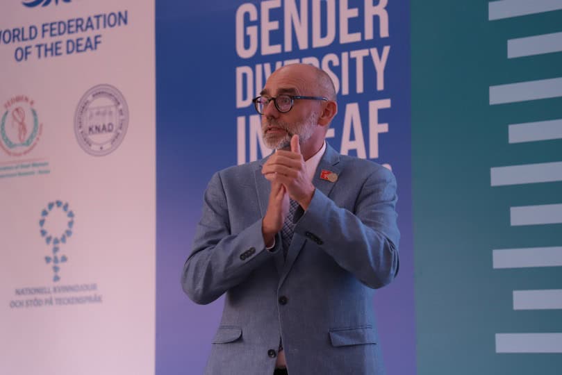 A man in a suit signs in front of banners about the World Federation of the Deaf and gender diversity at an event.