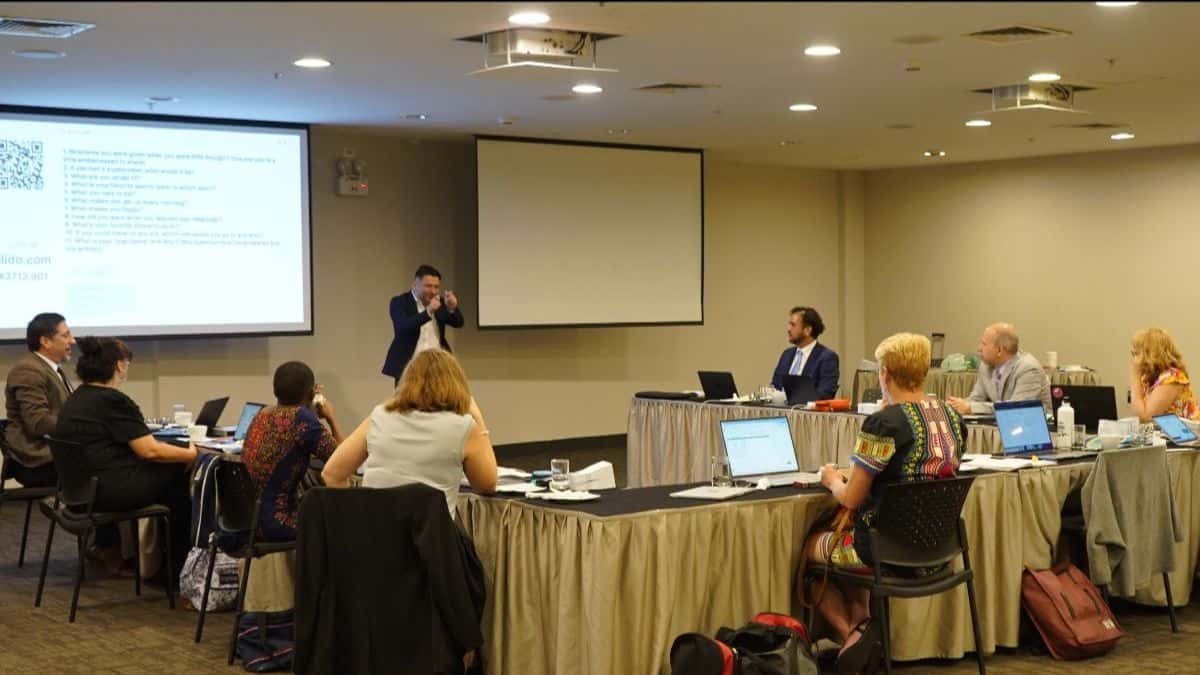 A group of people sit at tables in a conference room, listening to a presenter standing at the front near a screen displaying text. Laptops and notebooks are on the tables.