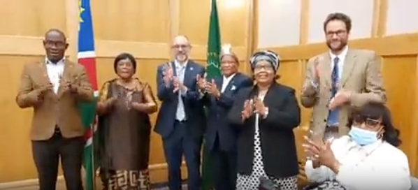 A group of seven people standing and clapping in a room with wooden walls, with a Namibian flag visible in the background.