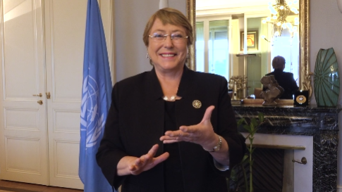 A person wearing a black blazer stands, gesturing with one hand. They are indoors, with a decorative mirror and a flag visible in the background.