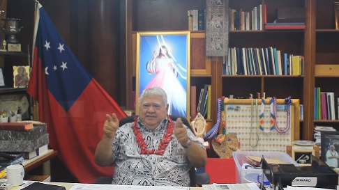 Person sitting at a desk in an office with a national flag and religious image in the background, gesturing with hands. Shelves filled with books and office supplies are visible.
