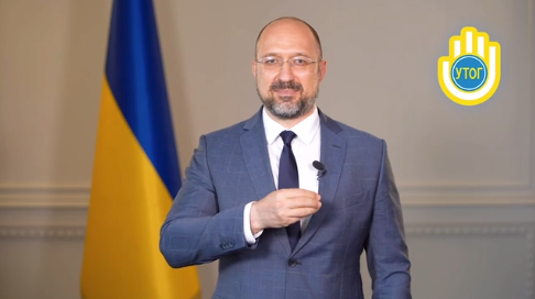 A man in a suit stands in front of a Ukrainian flag, gesturing, with a blue and yellow emblem on the upper right.