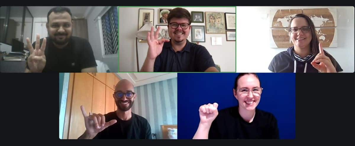 Five people in a video call, each holding one hand up in various gestures, smiling.