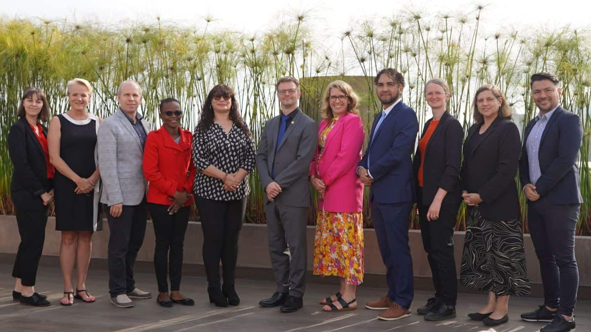 Group of ten people standing side by side outdoors on a patio with tall plants in the background.