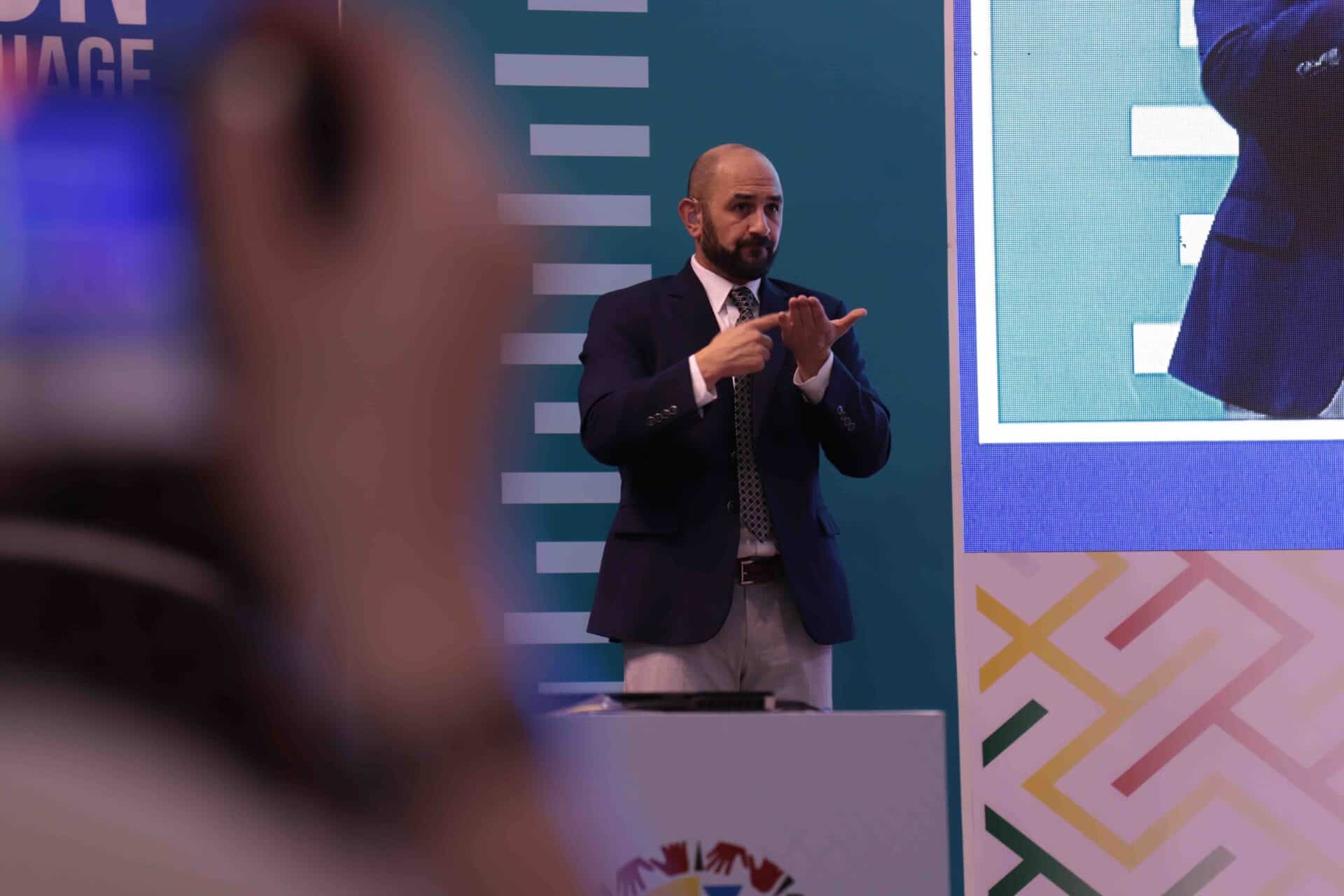 A man in a suit uses sign language on stage at an event, with a partial view of a sign language symbol and a large screen in the background.