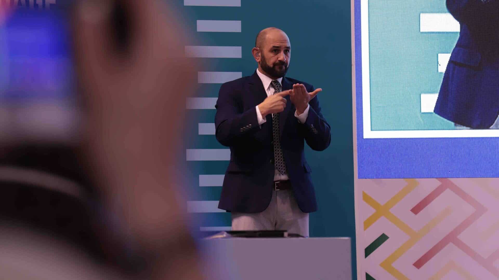 A man in a suit uses sign language on stage at an event, with a partial view of a sign language symbol and a large screen in the background.