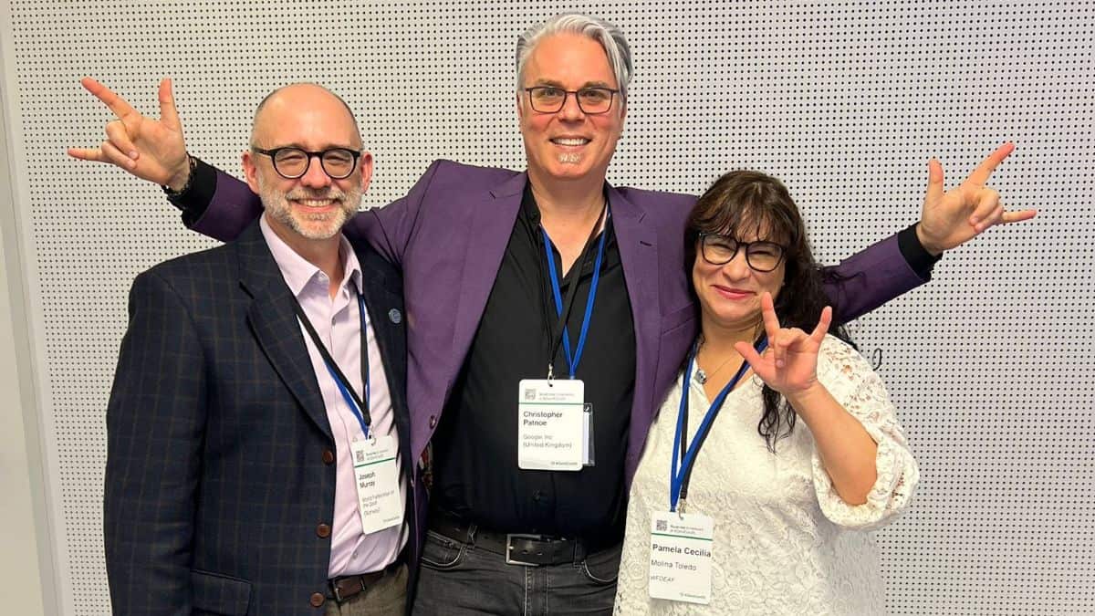 Three people posing happily in front of a white perforated wall, smiling and making hand gestures. All are wearing name tags and lanyards.
