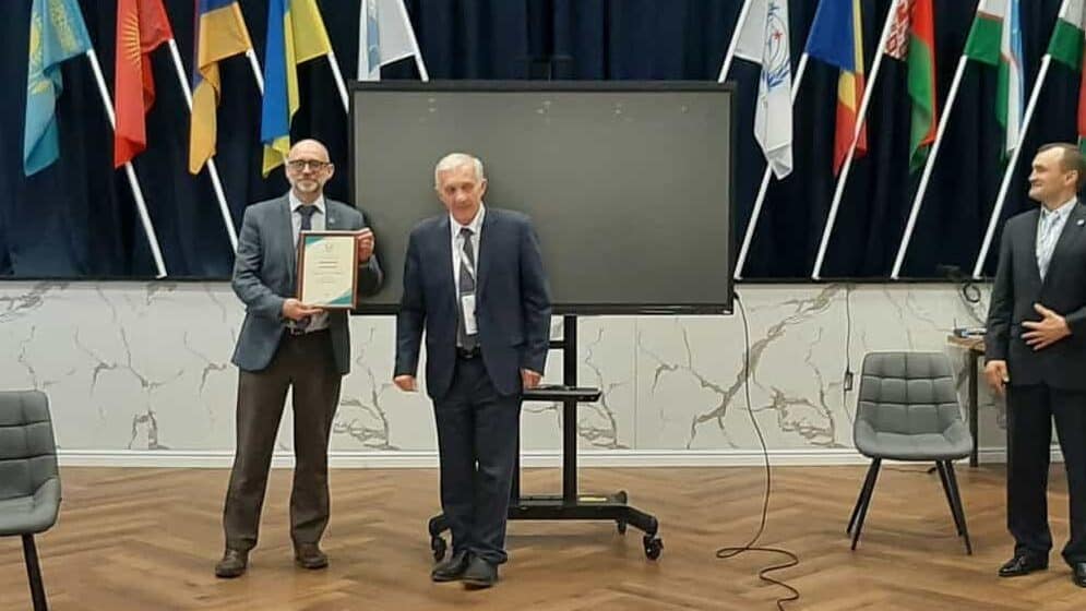 A group of people in business attire stand in a conference room with a display of international flags; two men pose with a certificate at the centre of the room.