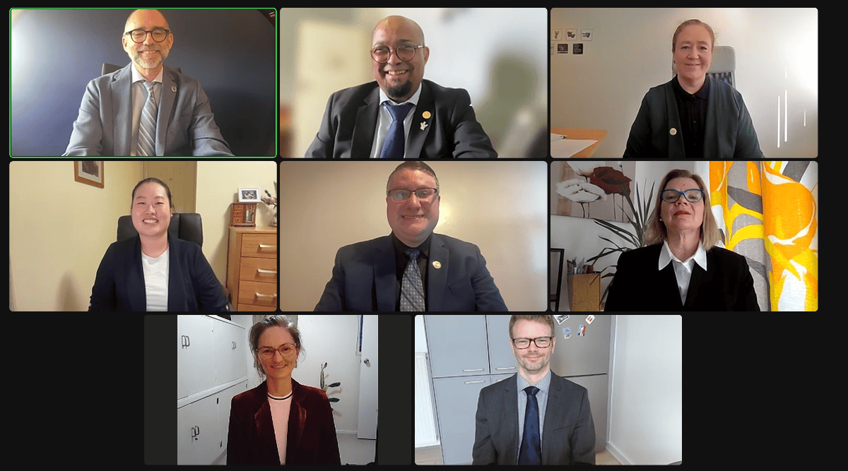 A virtual meeting with eight people, each in their own frame, smiling and wearing formal attire. A plant and artwork are visible in some frames.