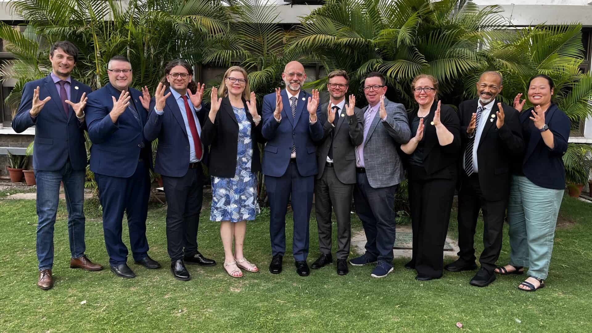Ten people in formal attire stand side by side outdoors on grass, smiling and making the Vulcan salute gesture with their hands. Lush green plants are in the background.