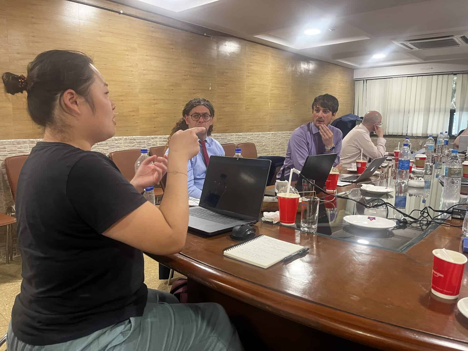 Woman speaking to two men during a meeting in a meeting room, with laptops, notebooks, and drinks on the table.