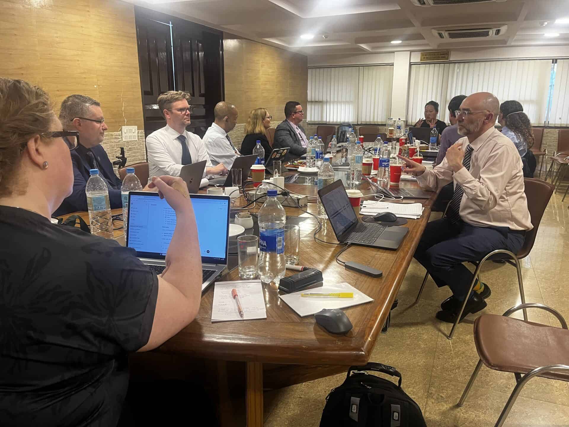 A group of people in business attire sit around a conference table with laptops, notebooks, and drinks, engaged in discussion in a well-lit meeting room.
