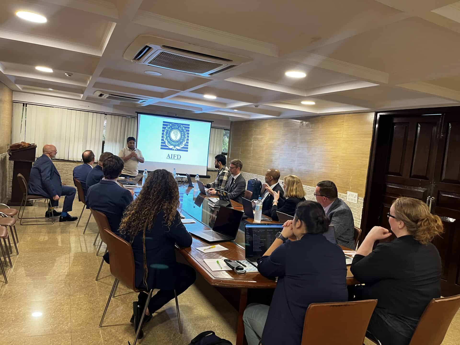 A group of people sit around a conference table watching a person present slides on a screen in a meeting room.