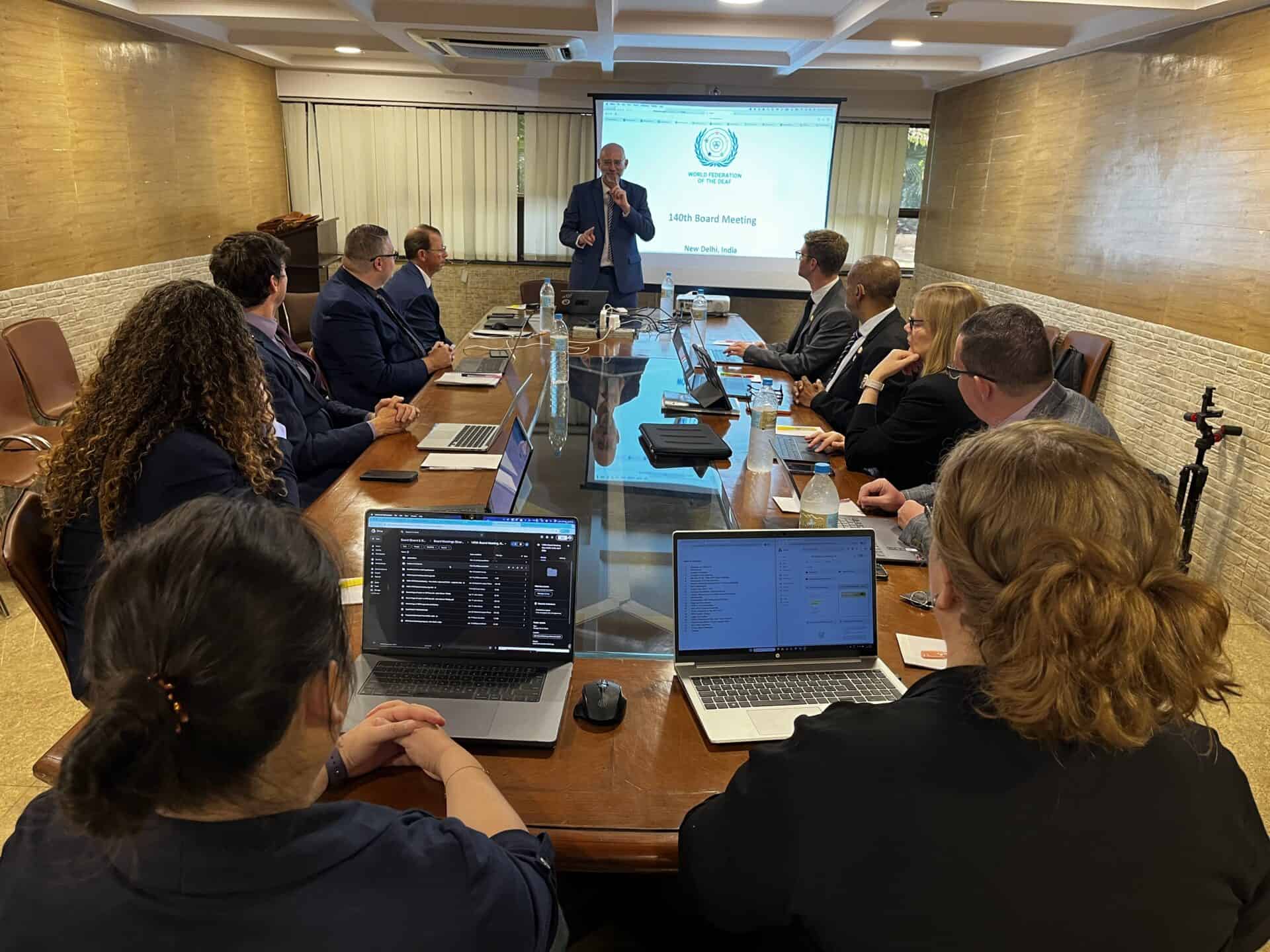 A group of people sit around a conference table with laptops, listening to a speaker presenting slides at a board meeting.