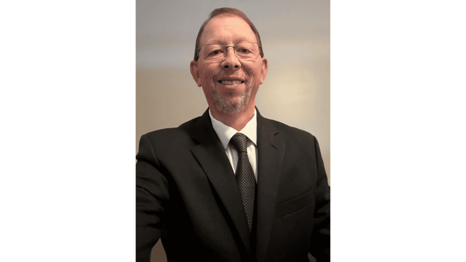 A man wearing glasses, a black suit, white shirt, and black tie stands in front of a plain beige background, looking at the camera and smiling.