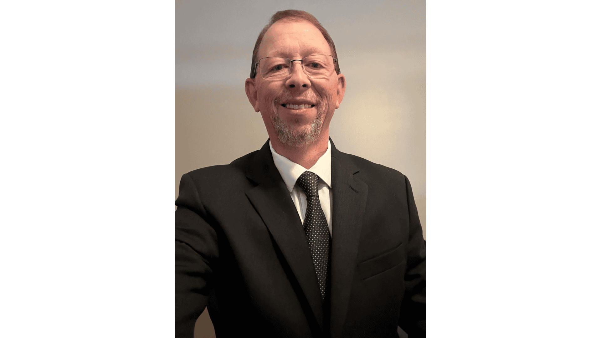 A man wearing glasses, a black suit, white shirt, and black tie stands in front of a plain beige background, looking at the camera and smiling.