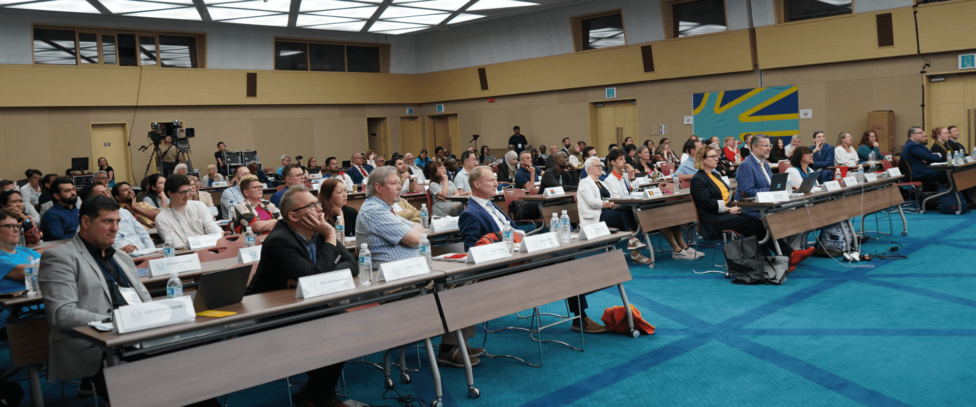 A large group of people sits at tables in a conference room, listening to a presentation. Name tags, microphones, and water bottles are visible on the tables.