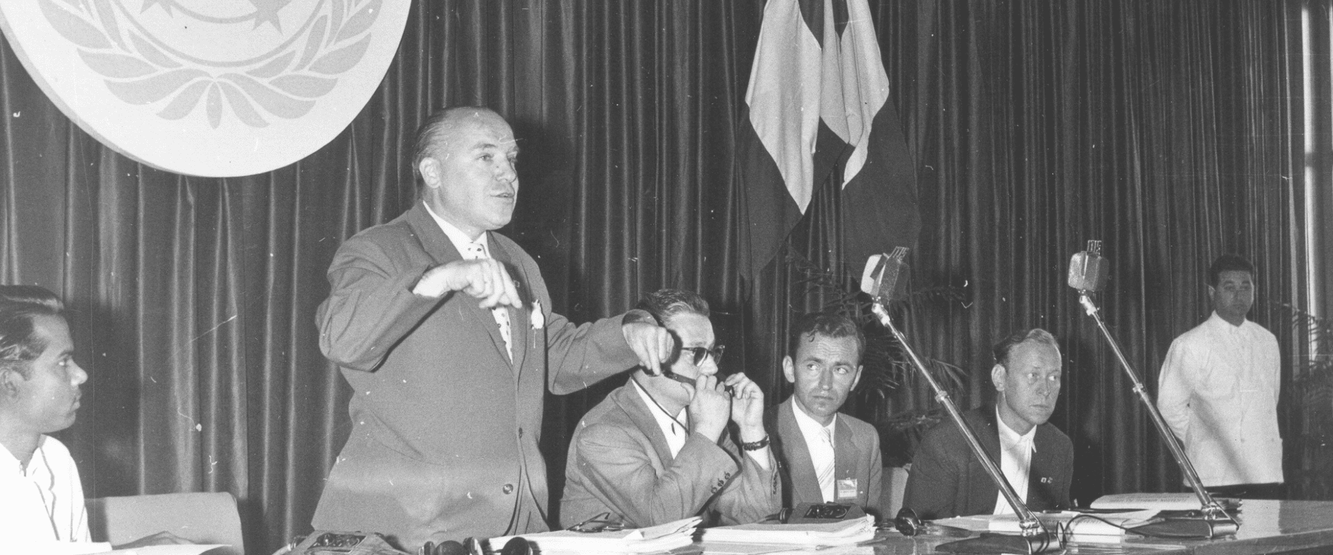 A man stands speaking at a table with several seated men and microphones, against a backdrop of flags and a large emblem.