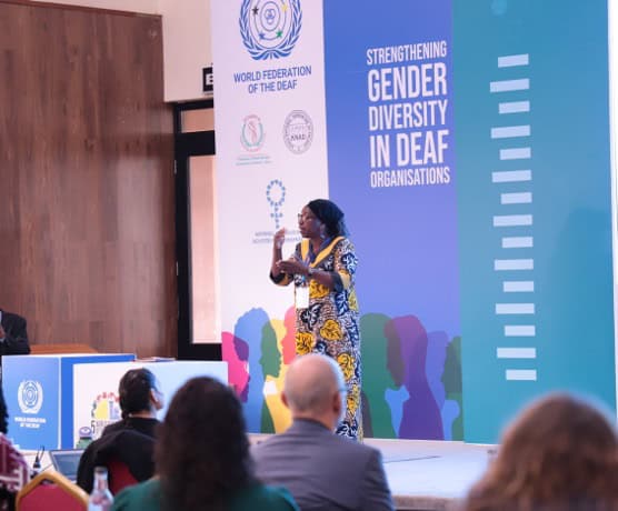 A woman speaks on stage at a conference focused on strengthening gender diversity in deaf organizations, with attendees seated in the audience.